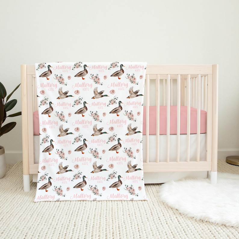 May include: A white baby blanket with a repeating pattern of ducks, pink flowers, and the name "Mallory Kate" in a cursive font. The blanket is draped over a crib with a pink sheet.
