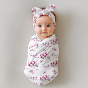May include: A white swaddle blanket with a floral pattern and the name "Abigail Emerson" repeated throughout. A baby is wrapped in the blanket and is wearing a white headband with a bow and the name "Abigail" written on it.