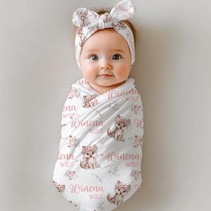 May include: A baby wrapped in a white swaddle blanket and wearing a matching headband. The blanket and headband feature a repeating pattern of a fox with a floral crown, pink flowers, and the text "Winona Wolf".