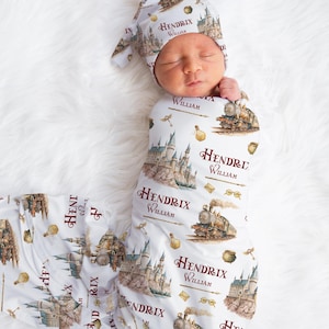 May include: A newborn baby wrapped in a white swaddle and matching cap, both adorned with a whimsical print featuring a castle, train, and the name "Hendrix William" in burgundy. The baby is nestled on a fluffy white surface.
