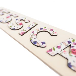 May include: Wooden name puzzle spelling out the name "Jessica". The letters are decorated with a floral pattern in shades of pink, purple, and white. The puzzle pieces are on a light-colored wooden base.