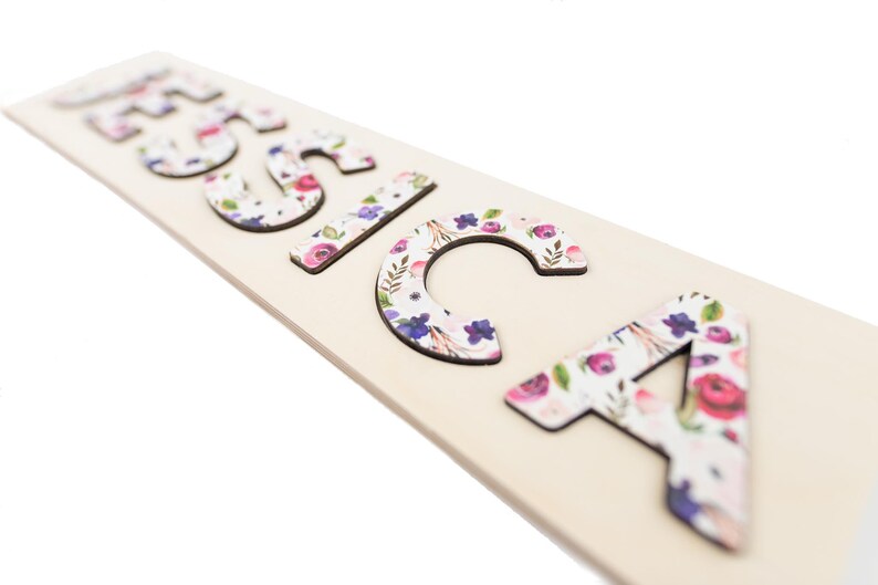 May include: Wooden name puzzle spelling out the name "Jessica". The letters are decorated with a floral pattern in shades of pink, purple, and white. The puzzle pieces are on a light-colored wooden base.