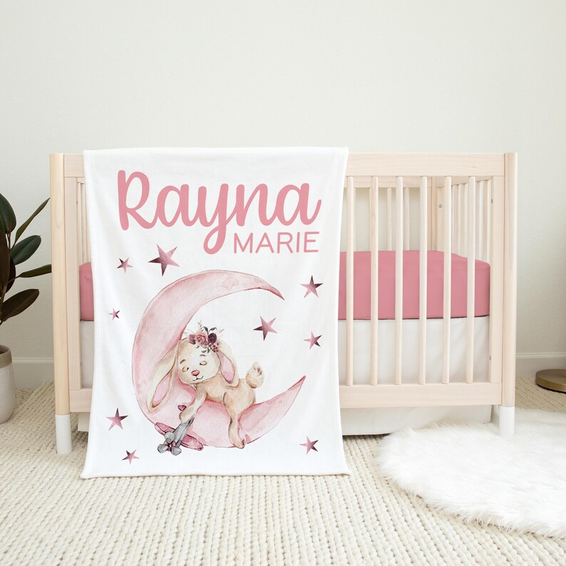 May include: A white baby blanket with a pink bunny rabbit sleeping on a pink crescent moon. The blanket has pink stars and the name "Rayna Marie" written in pink.