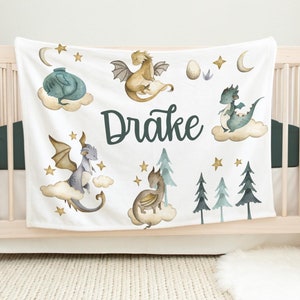 May include: White baby blanket with a green, blue, and gold dragon theme. The blanket features five dragons, stars, clouds, and the name "Drake".