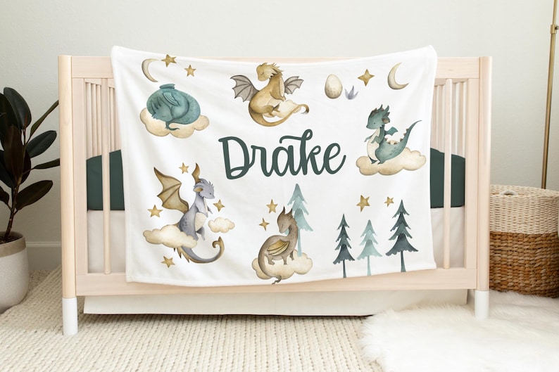 May include: White baby blanket with a green, blue, and gold dragon theme. The blanket features five dragons, stars, clouds, and the name "Drake".