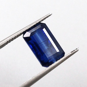 Puede incluir: Una gema rectangular de color azul oscuro sujeta con pinzas plateadas. La gema tiene un corte facetado con una superficie brillante y reflectante. El fondo es blanco liso, resaltando el color y la forma de la gema.