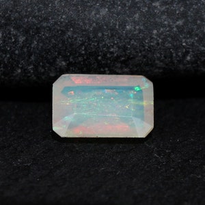 Może przedstawiać: Biały opal szlachetny o prostokątnym szlifie szmaragdowym. Opal ma zielono-czerwony, tęczowy połysk.