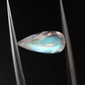 May include: A teardrop-shaped, iridescent gemstone with a pearlescent sheen. The gemstone is held by tweezers against a black background.