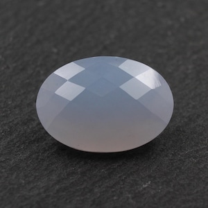 Puede incluir: Una piedra preciosa ovalada y facetada de color azul grisáceo translúcido. La piedra presenta una superficie lisa y pulida con facetas geométricas que reflejan la luz, creando un sutil juego de sombras sobre su forma redondeada y simétrica.
