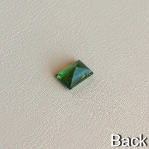 May include: A green gemstone with a rectangular shape, cut into a pyramid shape. The gemstone is on a white background.