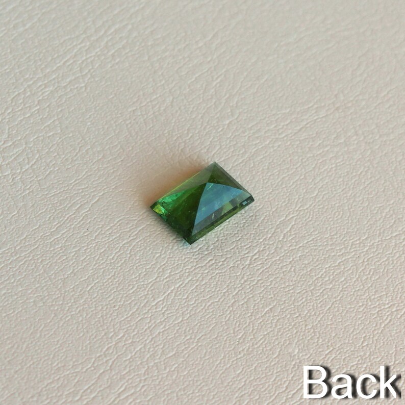 May include: A green gemstone with a rectangular shape, cut into a pyramid shape. The gemstone is on a white background.