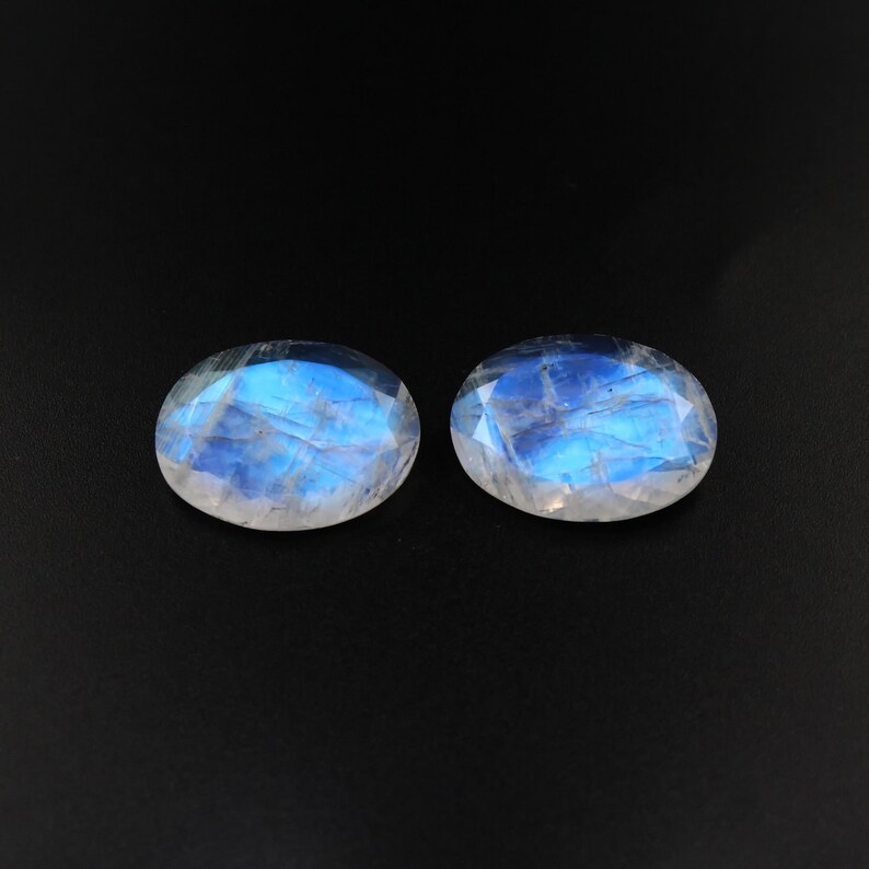 May include: Two oval-shaped moonstone gemstones with a blue iridescent sheen on a black background.