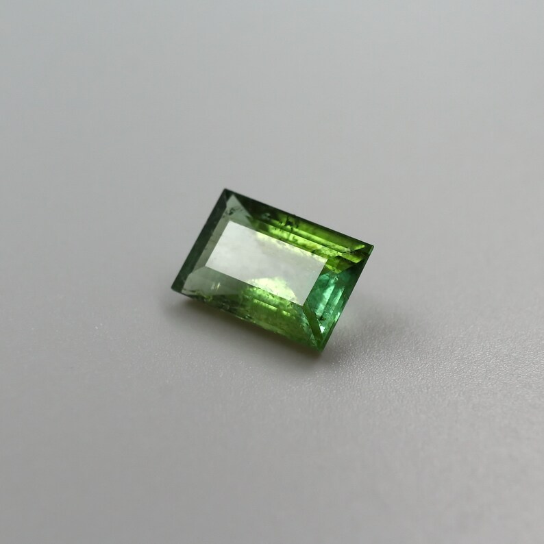 May include: A green and light green rectangular cut gemstone with facets.