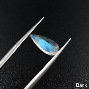 May include: A teardrop-shaped gemstone with a blue iridescent sheen, held by tweezers against a black background. The back side of the gemstone is visible.