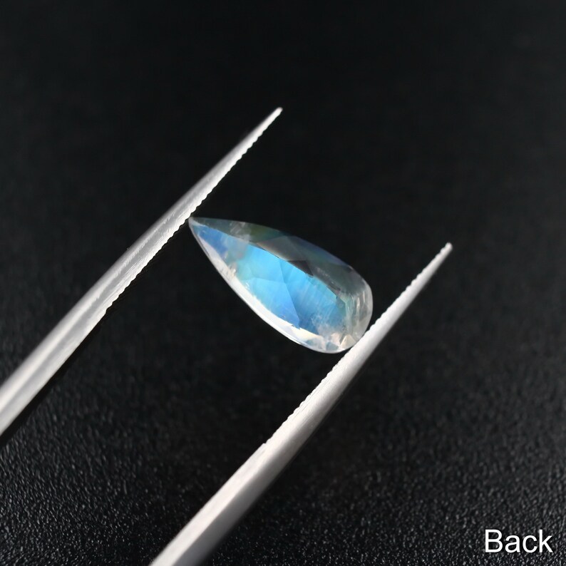 May include: A teardrop-shaped gemstone with a blue iridescent sheen, held by tweezers against a black background. The back side of the gemstone is visible.