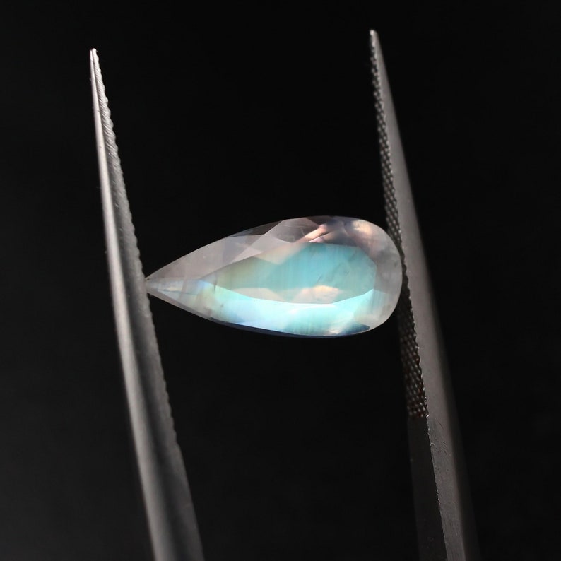May include: A teardrop-shaped, iridescent gemstone with a pearlescent sheen. The gemstone is held by tweezers against a black background.