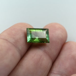 May include: A green gemstone with a rectangular cut, set in a bezel setting. The gemstone is a vibrant green color and has a high level of clarity.
