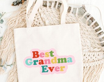 May include: A cream-colored tote bag with the text "Best Grandma Ever" in a colorful, retro font. The bag has two long handles and is made of a sturdy fabric. The background includes decorative elements.