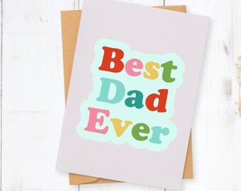 May include: A greeting card with the text "Best Dad Ever" in colorful bubble letters. The card is light purple and rests on a brown envelope. The background is a white wooden surface.