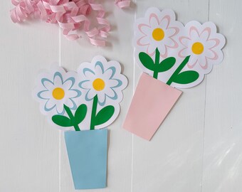May include: Two paper flower bouquets in blue and pink pots with white petals, yellow centers, and green stems. A pink ribbon curls in the upper left corner. The flowers are on a white wooden surface.