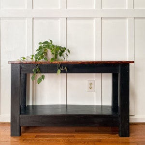 May include: A black console table with a wooden top and a lower shelf. The table is in front of a white wainscot wall and has a potted plant on top.