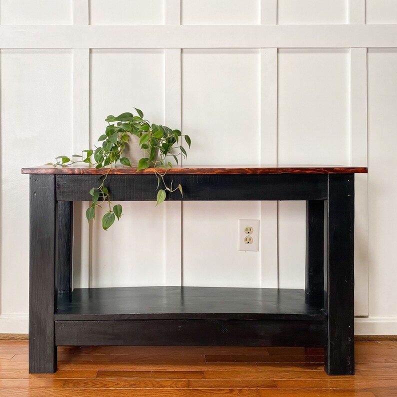 May include: A black console table with a wooden top and a lower shelf. The table is in front of a white wainscot wall and has a potted plant on top.