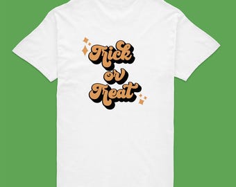May include: A white short-sleeved t-shirt featuring the text Trick or Treat in a stylized orange font with a black drop shadow. The design includes small four-pointed star accents in orange, positioned above and below the text on the shirt.