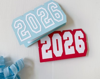 May include: Two graduation-themed cards with the year 2026 in white block letters. One card is light blue, the other is red. A blue ribbon is in the lower left corner.