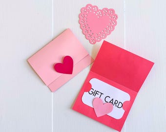 May include: Pink and red paper gift card holders with heart accents. One holder is closed with a red heart, the other is open with a gift card and a pink heart. A pink heart doily is also present. Text at the top reads: SVG • DXF • PNG • JPG.