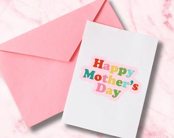 May include: A white greeting card with the text "Happy Mother's Day" in colorful bubble letters, accompanied by a pink envelope. The card and envelope are set against a pink marble background.