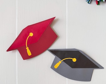 May include: Two graduation caps, one red and one gray, with yellow tassels. The red cap is on the left, and the gray cap is on the right. The caps are made of paper and are set against a white background.