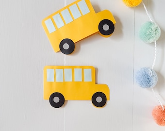 May include: Two yellow paper school bus cutouts with black wheels and light blue windows are displayed against a white background. A colorful pom-pom garland hangs on the right side, adding a playful touch.