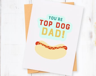 May include: A greeting card with a hot dog illustration and the text "YOU'RE TOP DOG DAD!" in a playful font. The card is white with a light brown envelope, set against a white wooden background.