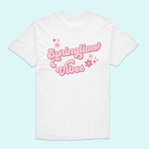 Puede incluir: Camiseta blanca con el texto "Springtime Vibes" en rosa, acompañada de flores rosas y formas de diamante. La camiseta tiene mangas cortas y cuello redondo, ideal para la primavera.