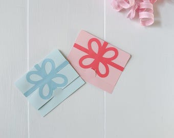 May include: Two gift cards, one light blue and one pink, each with a cut-out bow design. A pink ribbon curl is in the upper right corner. The cards are on a white wooden surface.