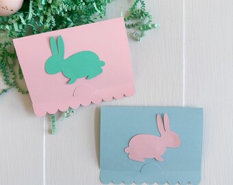 May include: Two Easter-themed paper crafts. One is light pink with a green bunny, the other is light blue with a pink bunny. Both have a scalloped edge at the bottom. The top of the image has the text "SVG • DXF • PNG • JPG".