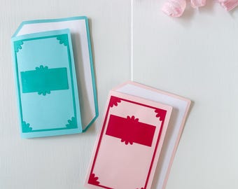 May include: Two decorative cards in the image. One is light blue with a teal accent, and the other is pink with a red accent. Both cards have a white backing and decorative corners. A pink ribbon is in the background.