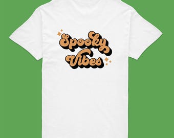 May include: A white short-sleeved t-shirt featuring the text Spooky Vibes in a tan, rounded, retro-style font with a black drop shadow. The design includes small four-pointed star accents in tan, positioned around the lettering on the shirt.