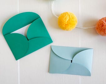 May include: Two paper envelopes, one teal and one light blue, are displayed on a white surface. The teal envelope is open, revealing a white interior. The image includes text at the top: SVG • DXF • PNG • JPG. A garland of yellow and orange pom-poms is in the background.
