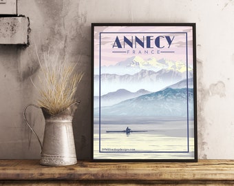 May include: A vintage travel poster design featuring a scenic view of Annecy, France. The poster has a light blue and pink colour scheme with a mountain range in the background and a lake in the foreground. The text "Annecy - France" is displayed in a bold, dark blue font.
