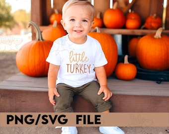 Puede incluir: Una camiseta blanca con el texto "little turkey" en letras doradas y naranjas. Un niño pequeño lleva la camiseta y está sentado en una plataforma de madera con calabazas en el fondo.