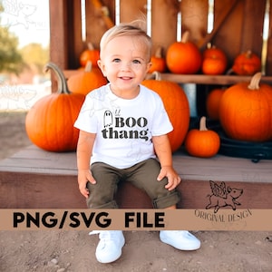May include: A young child wearing a white t-shirt with the text "lil BOO thang" and a ghost graphic, paired with olive green pants and white sneakers. The background features several orange pumpkins, suggesting a Halloween or autumn theme.
