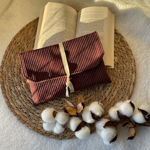 Op de afbeelding: Een bruine corduroy clutch met een wit lint en een kwast. De clutch ligt op een geweven stro mat met wattenbolletjes en een gedroogde bloem.