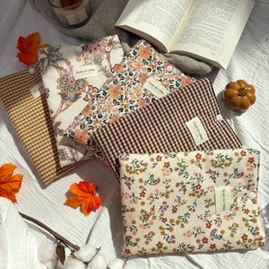 Könnte beinhalten: Vier rechteckige Buchhüllen aus Stoff mit Druckknopfverschluss. Eine hat ein braunes Vichy-Muster, eine einen cremefarbenen Blumendruck, eine ein dichtes orangefarbenes Blumenmuster und eine ein braun-weißes Vichy-Karo. Jede hat ein Etikett mit Fleurs de coton.