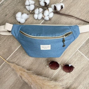 Puede incluir: Una riñonera de mezclilla azul claro con una cremallera blanca y una correa crema. La riñonera tiene una pequeña etiqueta marrón que dice "Fleurs de coton".