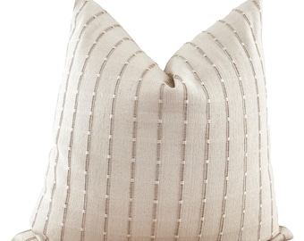 May include: A square throw pillow featuring a light beige fabric base with a vertical pattern of thin, dashed brown lines and small white stitch accents. The pillow has a textured, woven appearance with a structured shape and pointed top corners.