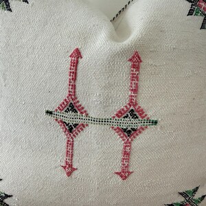 May include: A white cushion with an embroidered design. The embroidery features a repeating pattern of pink arrows and diamond shapes, with black and green accents. The cushion has a textured surface.