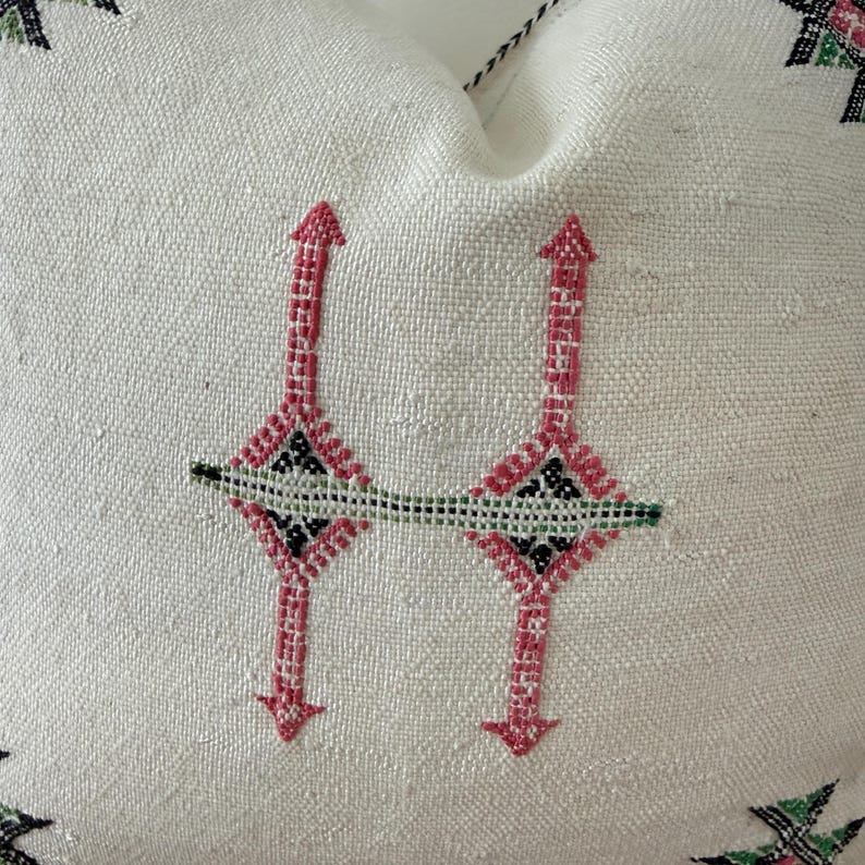 May include: A white cushion with an embroidered design. The embroidery features a repeating pattern of pink arrows and diamond shapes, with black and green accents. The cushion has a textured surface.