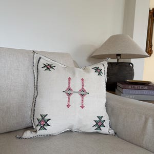 May include: A square, cream-coloured cushion featuring a geometric pattern in pink, black, and green. The cushion has a black stitched border and is placed on a beige sofa. Home decor.