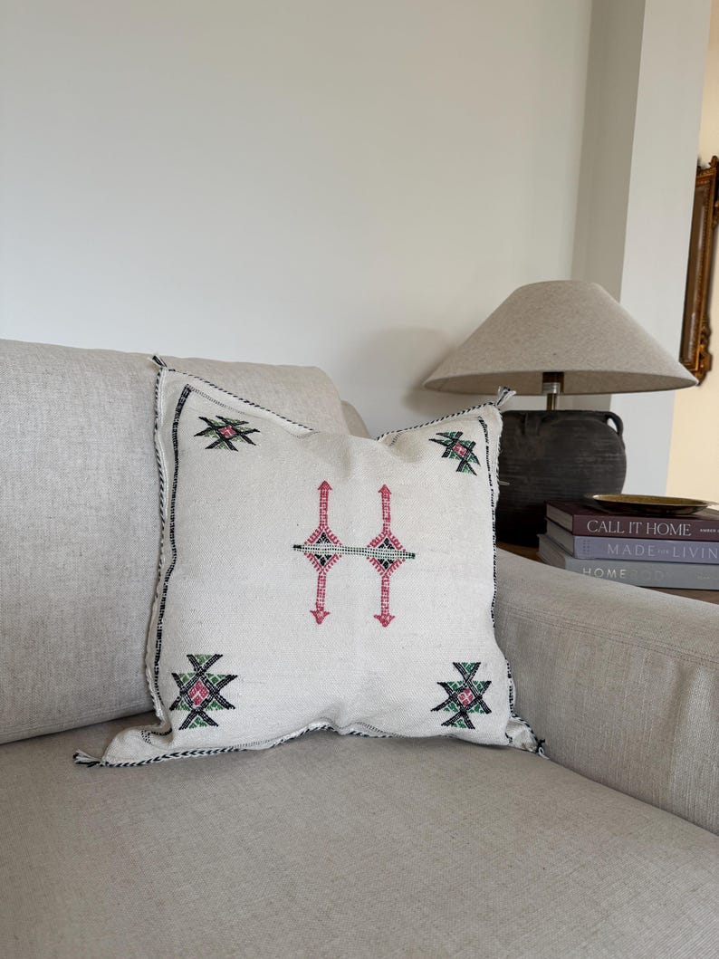 May include: A square, cream-coloured cushion featuring a geometric pattern in pink, black, and green. The cushion has a black stitched border and is placed on a beige sofa. Home decor.